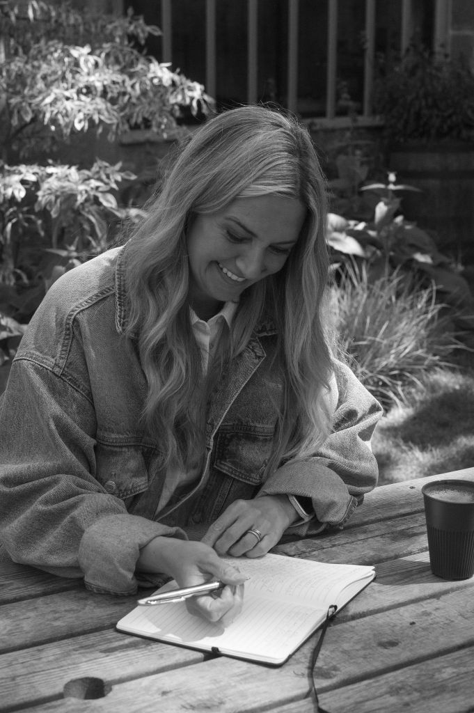 Woman sitting at an outside table writing in a notebook. There is a coffee cup next to her and some plants behind her. She is smiling.