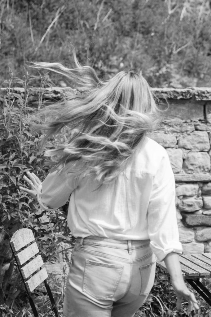 Black and white image of woman skipping outside near a table and chair. Long hair swaying in the wind.