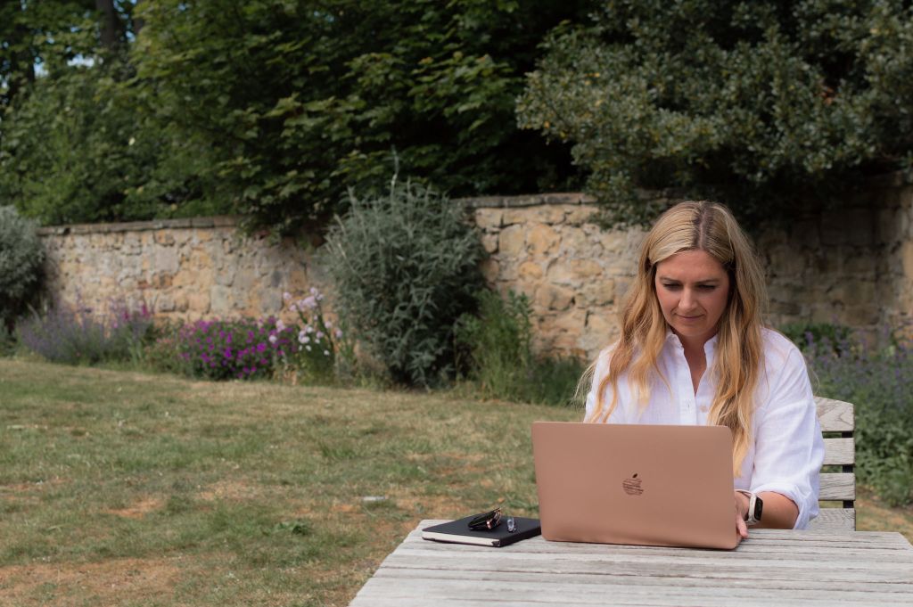 Copywriter sitting at a table in walled garden. She is looking intently at the open laptop on the table. Beside her are a book and a pair of glasses.