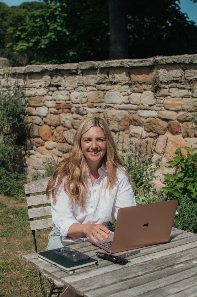 Woman sitting outside at a table looking at the camera smiling. On the table is an open laptop, a pile of books, a mobile phone and sunglasses. 