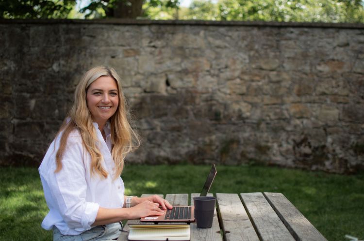 Woman sitting at picnic table with laptop open. She is looking at the camera and smiling.